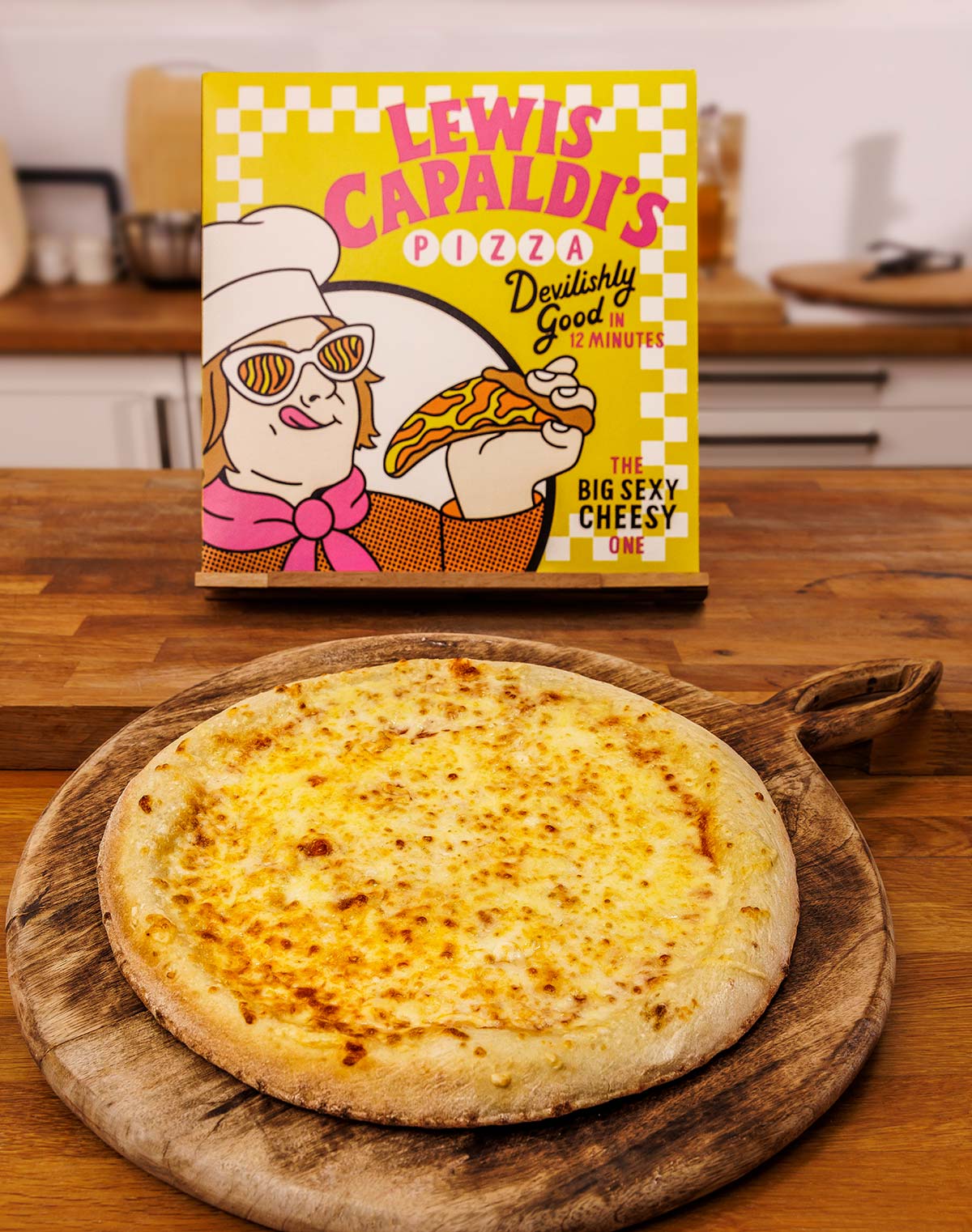 Photo of a cheese pizza on a wooden serving board, with a pizza box behind, branded 'Lewis Capaldi's Pizza'