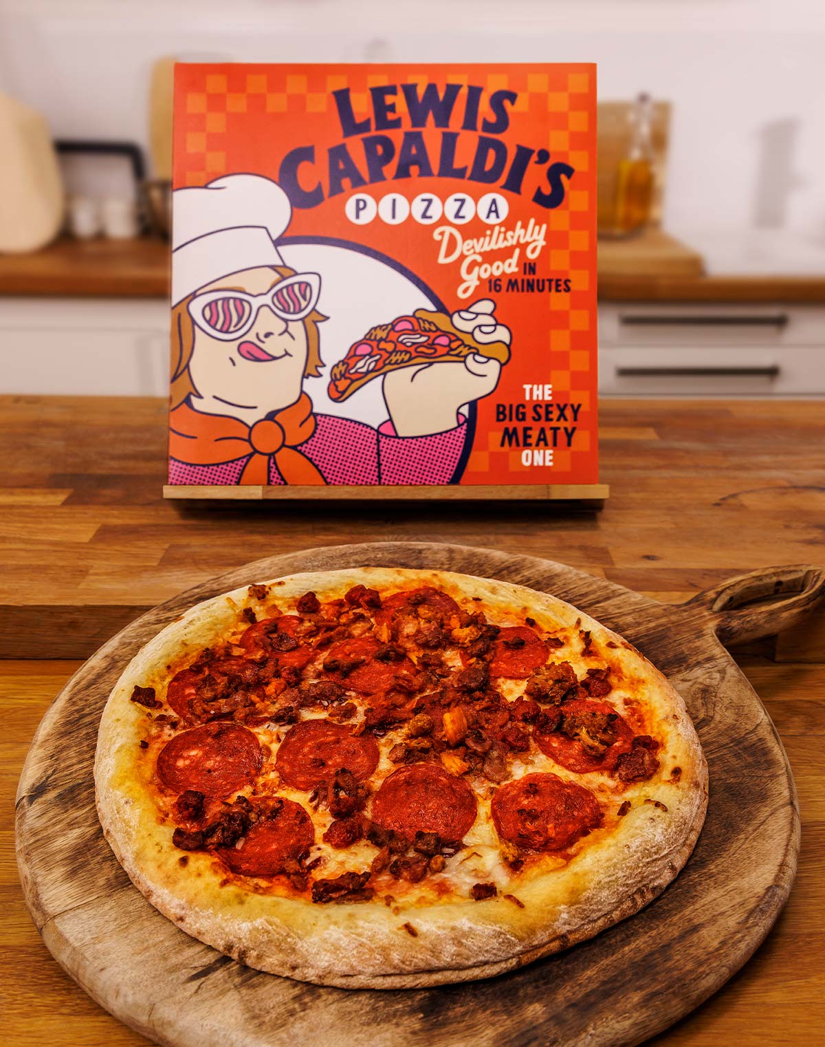 Photo of a meaty pizza on a wooden serving board, with a pizza box behind, branded 'Lewis Capaldi's Pizza'
