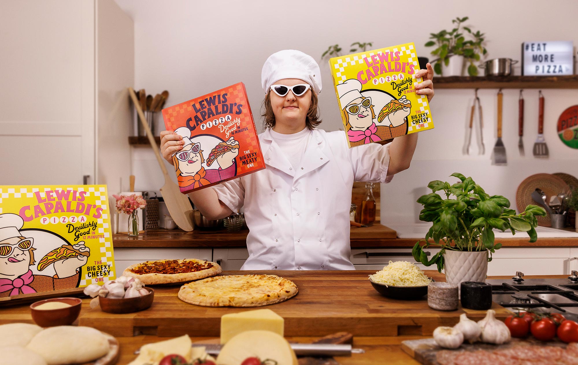 Photograph of Lewis Capaldi in a kitchen, wearing chef whites, holding two pizza boxes and standing infront of a table with pizzas, garlic, cheese and herbs.